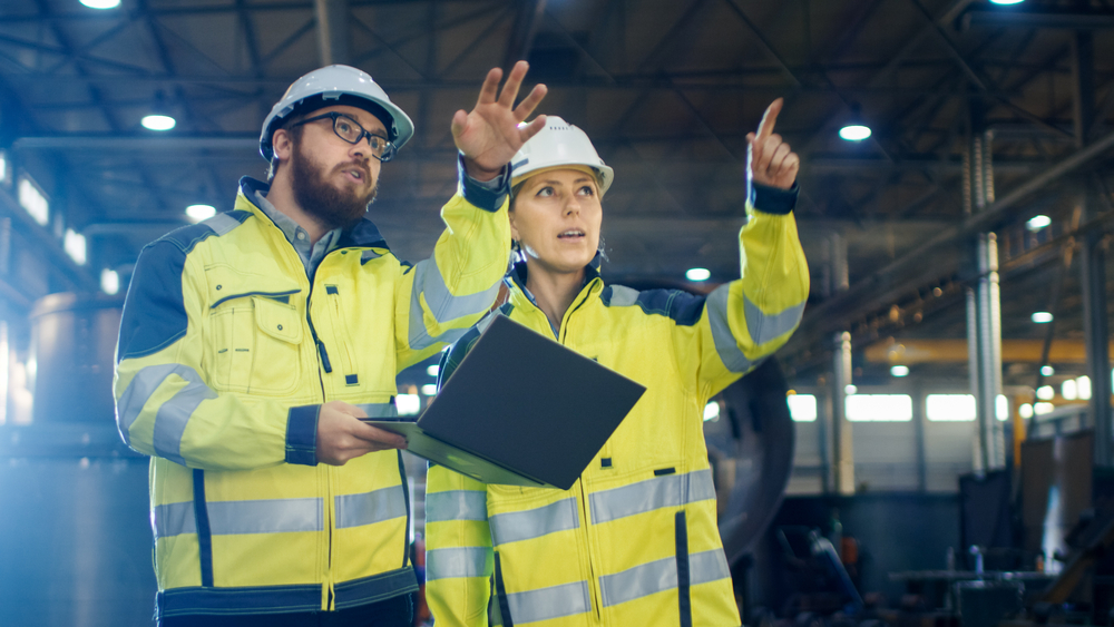 Male and Female Industrial Engineers Having Discussion and Use Laptop While Walking Through Heavy Industry Manufacturing Factory. They Wear Hard Hats and Safety Jackets.